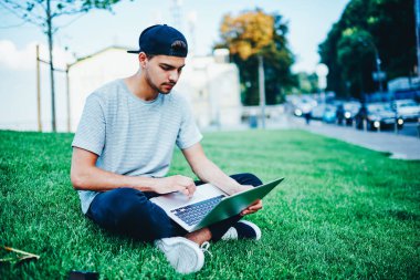 Dizüstü bilgisayar ile yeşil çimenlerin üzerinde oturan ve posta yoluyla ücretsiz 4 g kontrol gündelik kıyafeti dalgın öğrenci Internet Üniversitesi topraklarında. Netbook üzerinde USB'yi trendy şapkalı erkek blogger