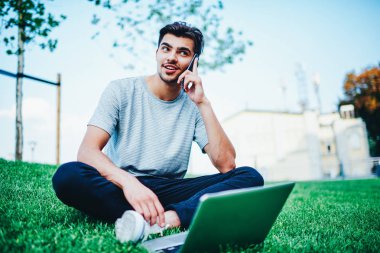 Pozitif hipster adam kampüs içinde modern dizüstü bilgisayar ile yeşil çimenlerin üzerinde otururken arkadaşıyla Smartphone aygıtında bahsediyoruz. Komik telefon cep üzerinde konuşmayı öğrenci