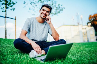 Neşeli hipster öğrenci komik telefon konuşma bir arkadaşım açık havada laptop ile üniversite topraklarında oturan smartphone sahip. Yeşil çimenlerin üzerinde hücresel dinlenme konuşan olumlu genç adam