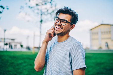 Optik gözlükler parkta dolaşmaya Smartphone cep telefonu ile en iyi arkadaşıyla konuşurken gülüyor içinde neşeli hipster blogger. Rahat t-shirt üzerinde cep komik konuşmayı olumlu genç adam