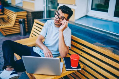 Dijital bilgisayar ile kentsel ortamda tahta bankta oturan şehre bağlıyken kameraya bakarak Eyewear zeki öğrenci portresi 4g ücretsiz internet. Dizüstü bilgisayar aygıtla oturan hipster adam