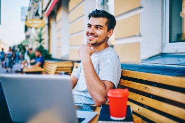 Kentsel ortamda dizüstü bilgisayar, tahta bankta oturan kablosuz 4 g için bağlıyken uzak görünen düşünceli gülümseyen genç adam Internet. Yakışıklı hipster blogger Caddesi üzerinde dinlenme netbook ile