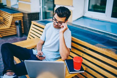 Kentsel ortamda dizüstü bilgisayar ile tahta bankta oturan smartphone takipçileri ile sohbet online optik gözlükler içinde dalgın hipster blogger bağlı 4 g ücretsiz internet