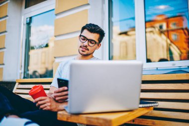 Modern dizüstü bilgisayar uzaktan çalışma 4 g ücretsiz bağlıyken bildirim Smartphone cep telefonu ile gönüllülük gözlük içinde erkek blogger şehir merkezinin kentsel ortamda tahta bankta oturmuş Internet