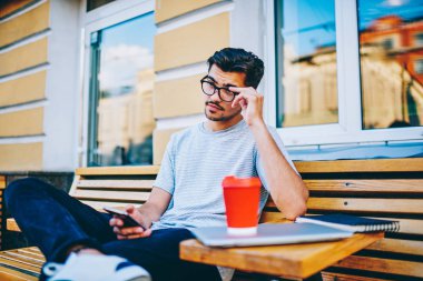 Optik gözlükler düz seyir ve posta smartphone ücretsiz 4 g internet bağlantısı ile kontrol ederken şehir merkezinde kahve ile tahta bankta oturan dalgın hipster blogger