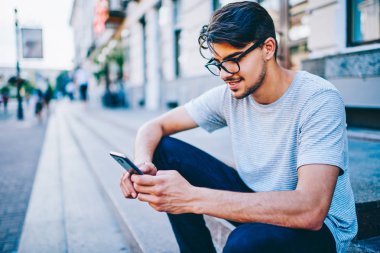 Neşeli erkek bogger takipçileri ile 4 g Smartphone cep telefonu ile internet sitesinde komik yorum okuma Internet. Pozitif hipster adam Eyewear iyi yeni şehir merkezinde oturan telefonu ile bildirim okuma