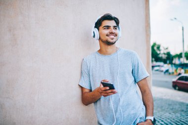 Neşeli hipster blogger dinleme elektronik müzik modern kulaklık açık havada kopyalama alanı alanı ile duvarının yakınında duran smartphone aygıt bağlı. Ses çalma listesinden zevk olumlu genç adam