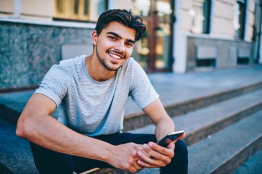 Ücretsiz yüksek hızda 4 g internet bağlantısı serbest zaman bir kentsel ortamda oturan smartphone aygıtında ile kendi web sayfasında yeni mesaj yayınlarken kameraya gülümseyen başarılı erkek blogger portresi