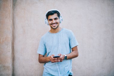 Kamera modern Kulaklık dinleme audiobook online iken gülümseyen mutlu hipster adam portresi smartphone ayakta promosyon arka plan üzerinde bağlı. Genç adam zevk müzik çalar