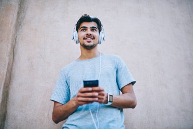 Olumlu erkek meloman görünümünü favori müzik zevk Smartphone cep telefonu ile 4 g üzerinden indirilen internet. Neşeli hipster blogger müzik şarkı yolu ile kulaklıklar açık havada duran Player'da radyo dinleme