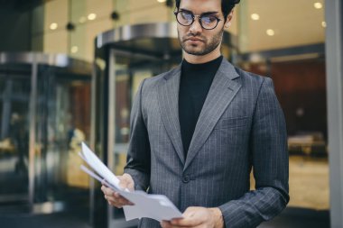 Başarılı bir iş adamı kırpılmış görüntü dikkatle kontrol finansal raporlar idari bina yakınındaki kentsel ortamda şık formal giyim giymiş. Dalgın gurur CEO'su okuma kağıt belgeleri