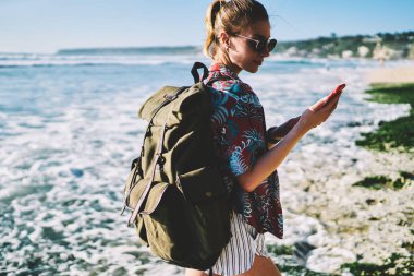 Hipster seyahat sırt çantası kıyı şeridinde yürürken Smartphone cep telefonu ile mesaj okuma okyanus su tarafından yıkarken yeni tropikal adada yaz macera sırasında yerler keşfetmek ile geziler kadın hayranı