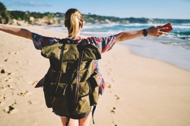 Güneşli kumlu sahili gezinme sırasında şık giyim eğlence içinde giyinmiş sarışın kadın kişi görüntü kırpılmış. Kadın turist yeşil sırt çantası ile arkadan görünüm zevk yaşam tatil sırasında
