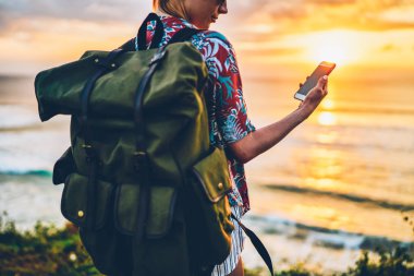 Cep telefonu bildirimini dusk okyanus yakın sırasında kontrol yeşil sırt çantası ile kadın turist resim kırpılmış. Arkadan görünüşü dışında yeniden oluştururken Web sitesinde telefon videoda izlerken hippi kız