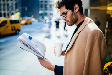 Trendy giyinmiş, işadamı Eyewear gazetede makaledeki bilgiler açık havada Manhattan üzerinde mola sırasında konsantre emin gitmek yakışıklı adam sabah haber kahve tutan basından okuma