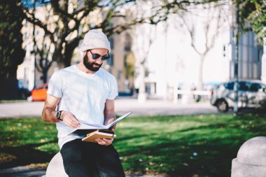İyi görünümlü olumlu akıllı erkek girişimci Sokağı'nda otururken çalışma programı Not Defteri'nden kontrol serin güneş gözlüğü. Ders kitabı dışında bilgi okuma mutlu hipster adam sakallı
