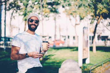 Gülümseyen, yarım uzunluk portre boş vaktinizi lezzetli içecek ve gadget ile şık güneş gözlüklü erkek kişi sakallı. Modern teknolojiyi kullanarak kentsel ayarı yeniden mutlu hipster adam