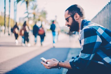 Genç erkek blogger yakışıklı şık kıyafet kısa mesaj için 4 g bağlı hücresel e-posta yoluyla göndermek için giyinmiş. Sakallı hippi adam açık havada ve holding sigara otururken online rezervasyon