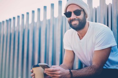 Yarım uzunluk portre rekreasyon Street içecek ve gadget ile zevk şık güneş gözlüğü gülümseyen yakışıklı. Pozitif hipster adam uygulama ağdan yükleme görüntüsünü kırpılmış