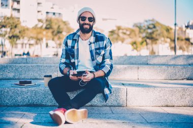 Yarım uzunlukta portre rekreasyon kulaklıklar ile en sevdiğiniz müzik ile zevk trendy güneş gözlüğü gülümseyen yakışıklı. Mutlu sakallı erkek kişi online podcast dinlerken kameraya bakıyor