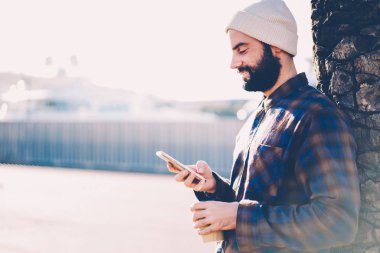 Yan görünüm gülümseyen, sakallı hippi lezzetli kahve elinde tutarak ve kendi blog smartphone ve 4 g kullanarak komik yorumlar okuma beyaz şapkalı adamı internet tanıtım alanı yakınında sokakta duran