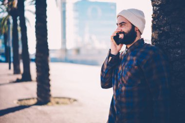 Arkadaş grubu smartphone aygıt üzerinde dolaşım söz şapka neşeli Genç sakallı adam yan görünüm. Cep telefonu açık havada kentsel ortamda ayakta iletişim rahat gömlekli olumlu hister adam