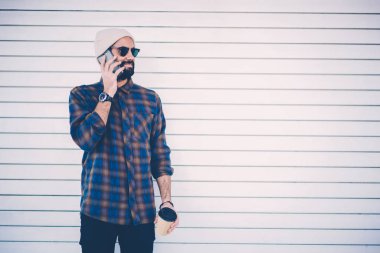 Hipster adam siyah güneş gözlüğü ve şık şapka tanıtım alanının yakınında promosyon arka plan smartphone aygıt ayakta arkadaşlarınızla konuşurken sakallı. Genç adam beyaz duvarının yakınında cep: çağrı