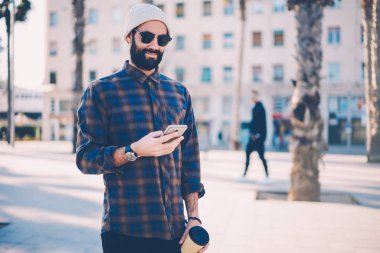 Gülümseyen genç adam şık güneş gözlüğü ele gider için sms mesajı açık havada kahve ile gezinme smartphone üzerinde okuma sakallı. Neşeli hipster adam sokakta yürürken cellualar e-posta kontrol şapkalı