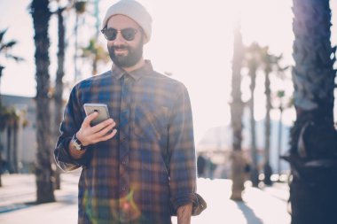 Güneş gözlüğü ve şık şapka urvan ortamda yürüyüş smartphone aygıt üzerindeki uygulama yükleme olumlu hipster adam. Sakallı öğrencinin cep sms mesajı okuma rahat kıyafetler giymiş