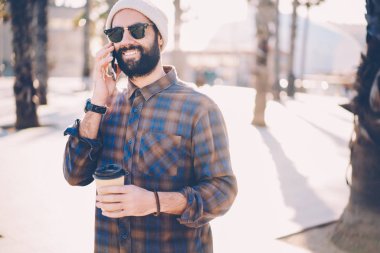 Mutlu sakallı adam lezzetli kahve ile sokakta el süre gezinme smartphone cep telefonu operatörü ile konuşurken şık casual kıyafet giymiş. Hipster adam telefonla iletişim güneş gözlüğü