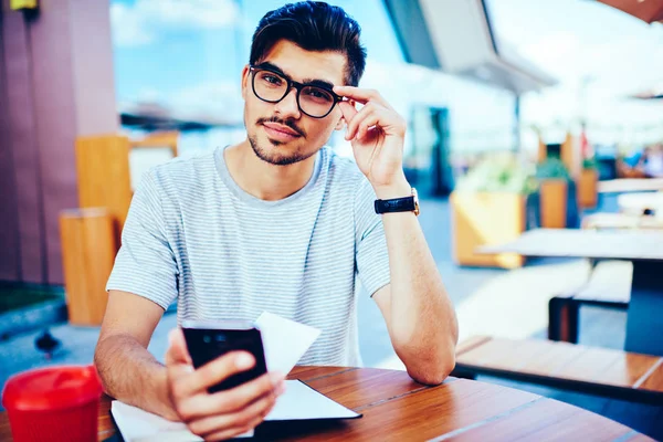 英俊的胡子时髦 Bloger 的肖像在时尚的眼镜看相机 而更新智能手机上的社交网络上的配置文件连接到免费的4G 互联网连接坐在户外 — 图库照片