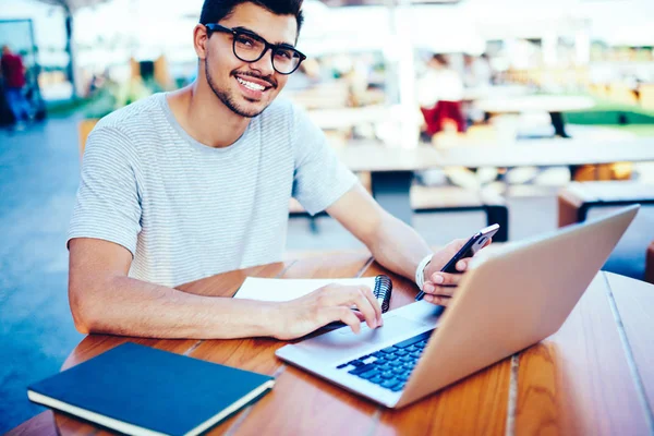 Portrait of successful male blogger in eyeglasses smiling at camera while synchronizes media files from smartphone to modern laptop computer using wireless internet connection for productive work