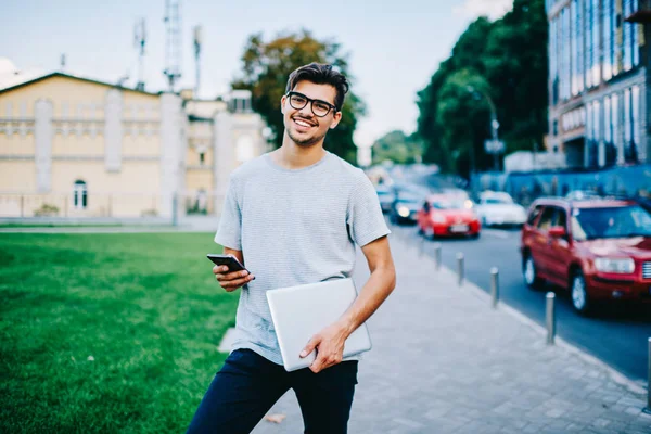 Portrait of successful casual dressed student in eyeglasses sending sms message to friend in social networks on modern smartphone connected to 4G internet walking in urban setting with laptop computer