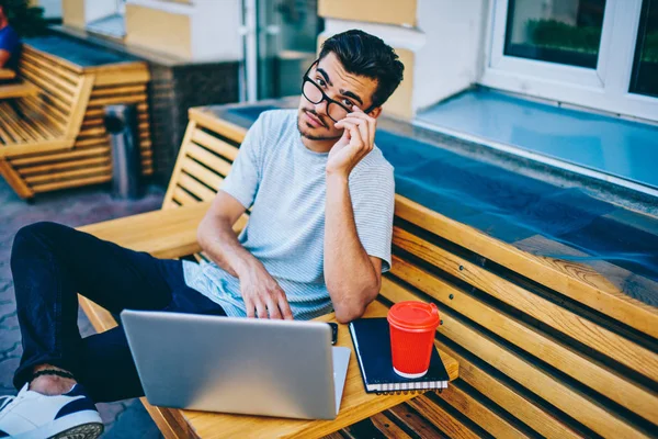 Dijital bilgisayar ile kentsel ortamda tahta bankta oturan şehre bağlıyken kameraya bakarak Eyewear zeki öğrenci portresi 4g ücretsiz internet. Dizüstü bilgisayar aygıtla oturan hipster adam