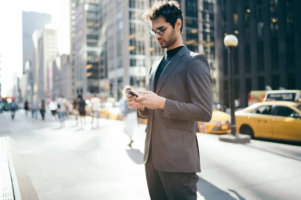 Intelligent executive director in formal wear standing in urban setting of Manhattan near office building and reading business news in application on smartphone via free 4G internet connection