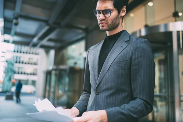 Disgruntled proud CEO dressed in formal wear annoyed with bad financial reports standing near office building.Dissatisfied businessman reading documentation with text information of data on street