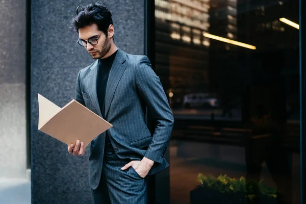 Fashionable young lawyer reading accounting reports strolling near office building.Pensive successful businessman in formal clothes checking file documentation walking in urban setting in downtown