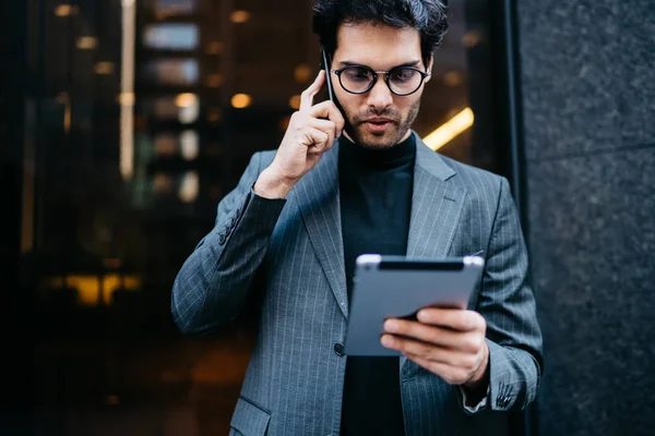 Successful young proud CEO with touch pad in hands reading message with business commercial offer received on mail while talking with partner on smartphone standing outdoors near office building