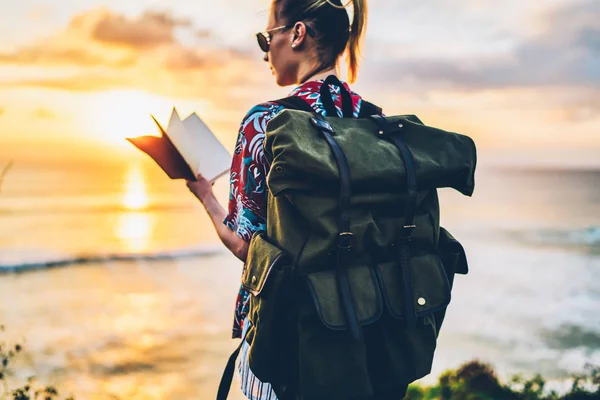 照片中的年轻女性人在笔记本上的图片检查笔记在休闲附近的海洋景观 背着绿色背包 站在海边的旅行者的背景色 — 图库照片