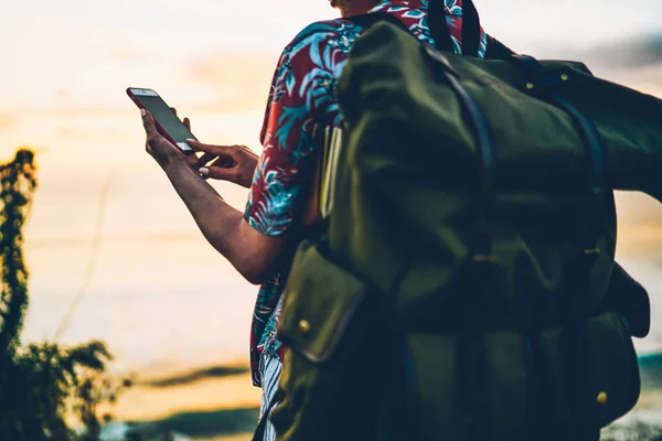 Back view of young woman tourist with big green rucksack sending video messages to friends for share impressions about vacation.Cropped image of female person enjoying recreating near to ocean