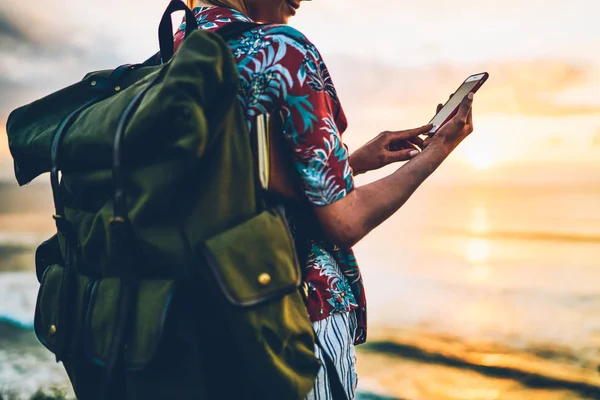 Kırpılmış görüntü alacakaranlıkta okyanus karşı boş zaman harcama yaparken büyük yeşil sırt çantası taşıyan kadın turist. Modern aygıtıyla iletişim için açık havada eğlence hippi kız Gezgin görünümünü geri