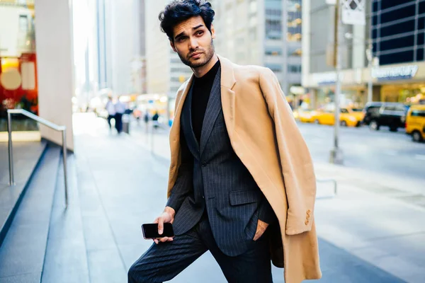 Handsome businessman dressed in elegant suit and coat looking away standing on urban setting, young confident entrepreneur dressed in fashion apparel holding smartphone spending time in downtow