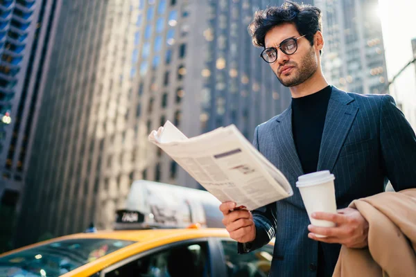 Confident male entrepreneur reading newspaper walking in business district of megalopolis holding coffee to go cup with copy space for name or logo,serious elegant man puzzled on article in pres