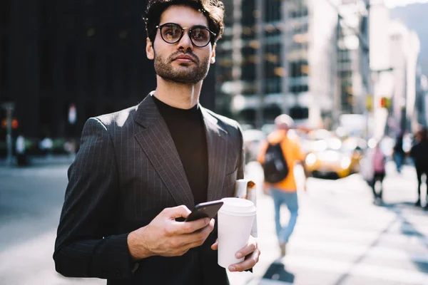 Handsome male in formal wear standing in New York downtown holding smartphone and coffee to go cup with copy space for brand name or logo, young businessman  sending text message via mobile on stree