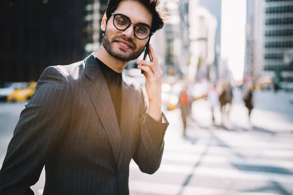 Smiling prosperous businessman in elegant wear satisfied with connection in roaming having mobile phone conversation on street,positive male in formal suit checking banking account calling to operato