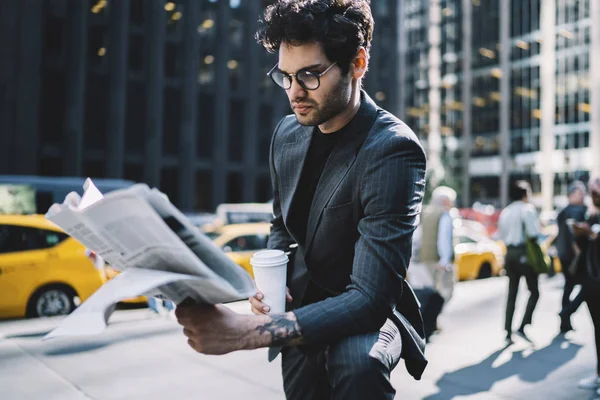 Handsome male professional in trendy formal outfit reading morning press with coffee to go in business district,confident man in elegant suit analyzing financial article in newspaper outdoors