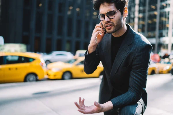 Annoyed professional manager annoyed during mobile phone conversation standing on avenue, angry businessman in suit worried about problem discusing solution through telephone talk gesturing in cit