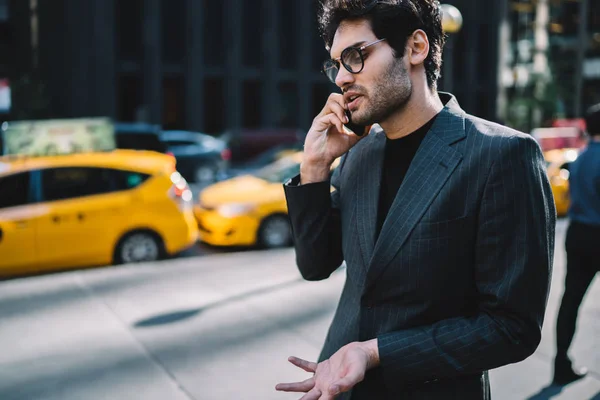 Professional man in trendy formal wear feeling angry solving problem through telephone conversation walking on street,emotional businessman in suit stressed discussing project talking on smartphone