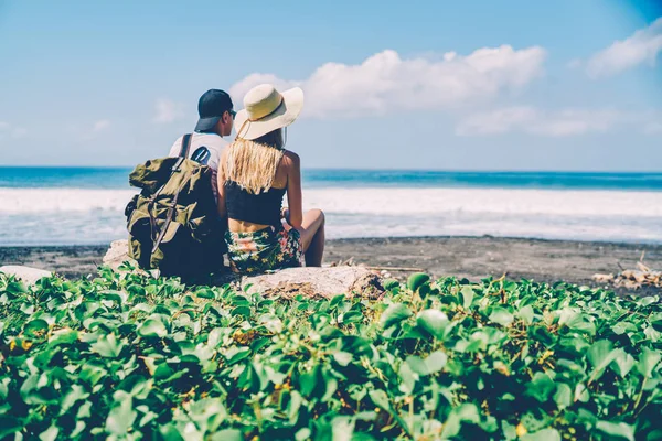Back view of romantic couple recreating on ocean shore looking at breathtaking scenery, hipster guy with touristic rucksack explore tropical island nature with girlfriend sitting near on seaside