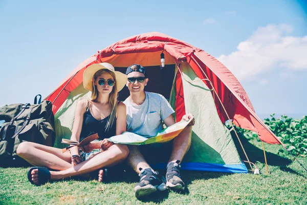Portrait of young male and female explorers sitting near camp with touristic equipment enjoying active weekends,couple of travelers camping on summer vacations looking at camera holding map